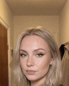 A silent reaction clip — she glances down at her phone, back up to the lens, and a suppressed smirk forms, mouth closed throughout