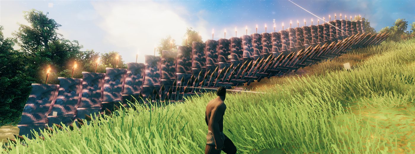 A long stone wall with stakes and torches, stepping up a grassy hill