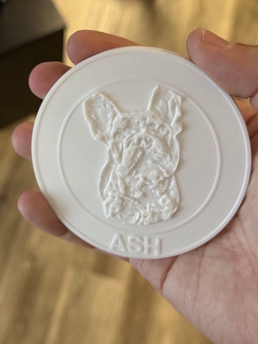 A 3D-printed coaster with a relief portrait of a French bulldog and the name ASH along the bottom edge, held in a hand