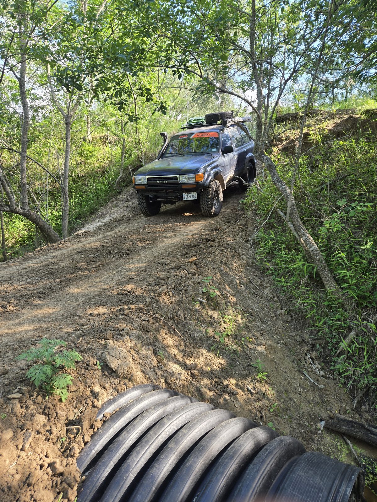 Toyota Land Cruiser descending a muddy trail past stacked culvert pipes