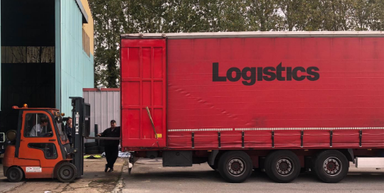 a forklift loading boxes onto a truck; the truck has the word ‘Logistics’ written across it