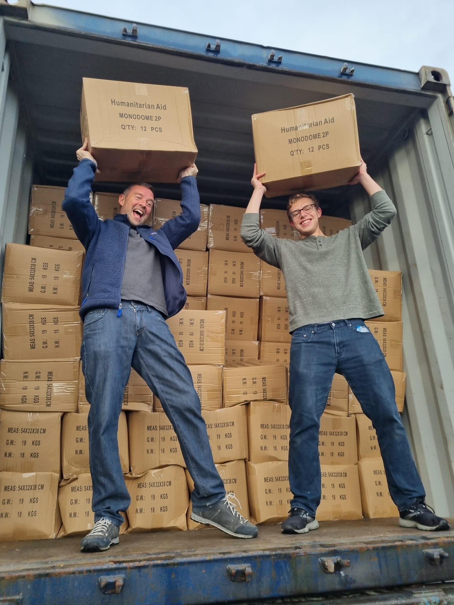 two volunteers standing in a truck full of boxes, holding two boxes (labelled ‘Humanitarian Aid’) of aid items above their head