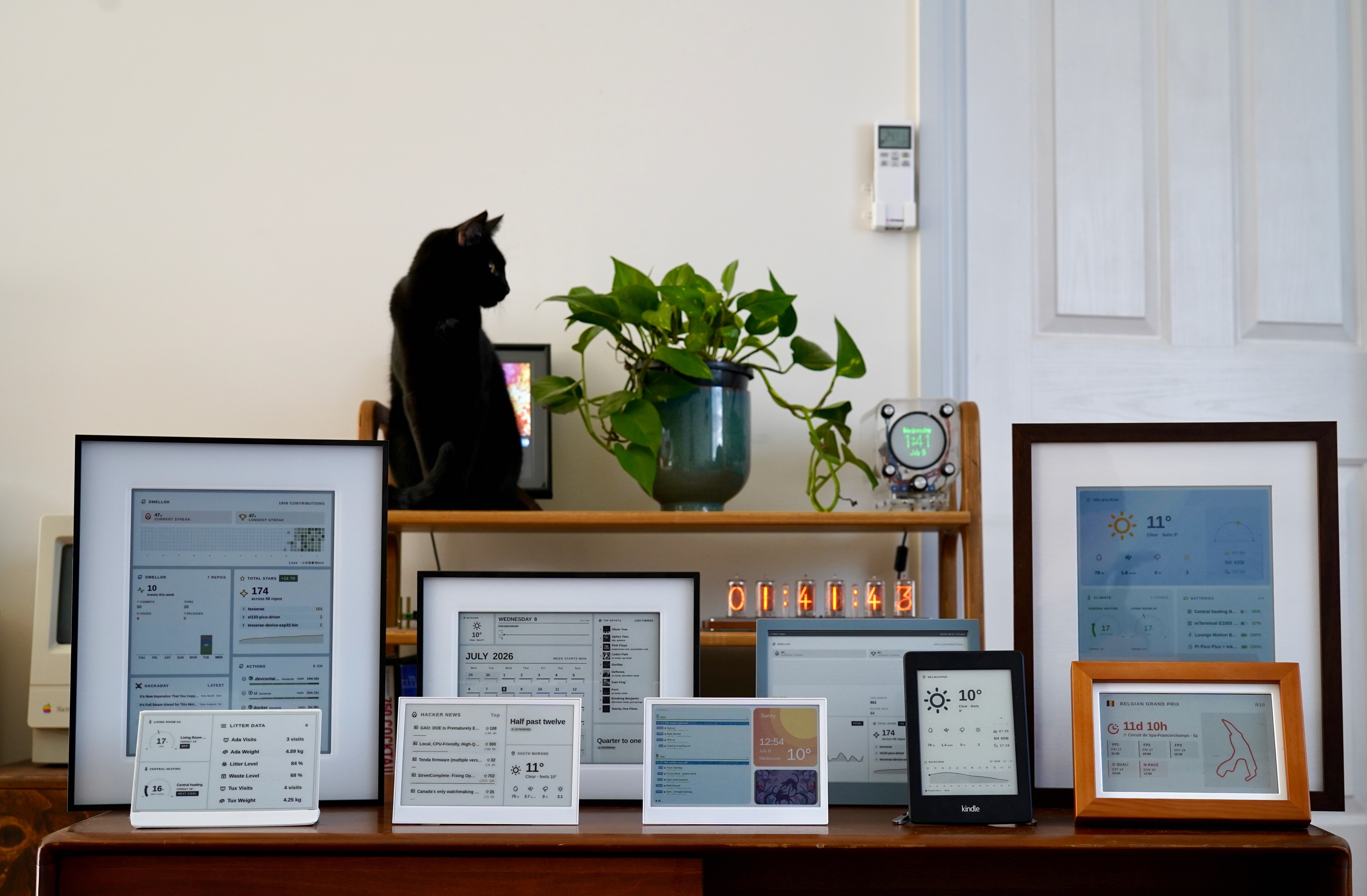 Nine e-ink panels on a wooden dresser, each running a different Tesserae dashboard, with a black cat in the background