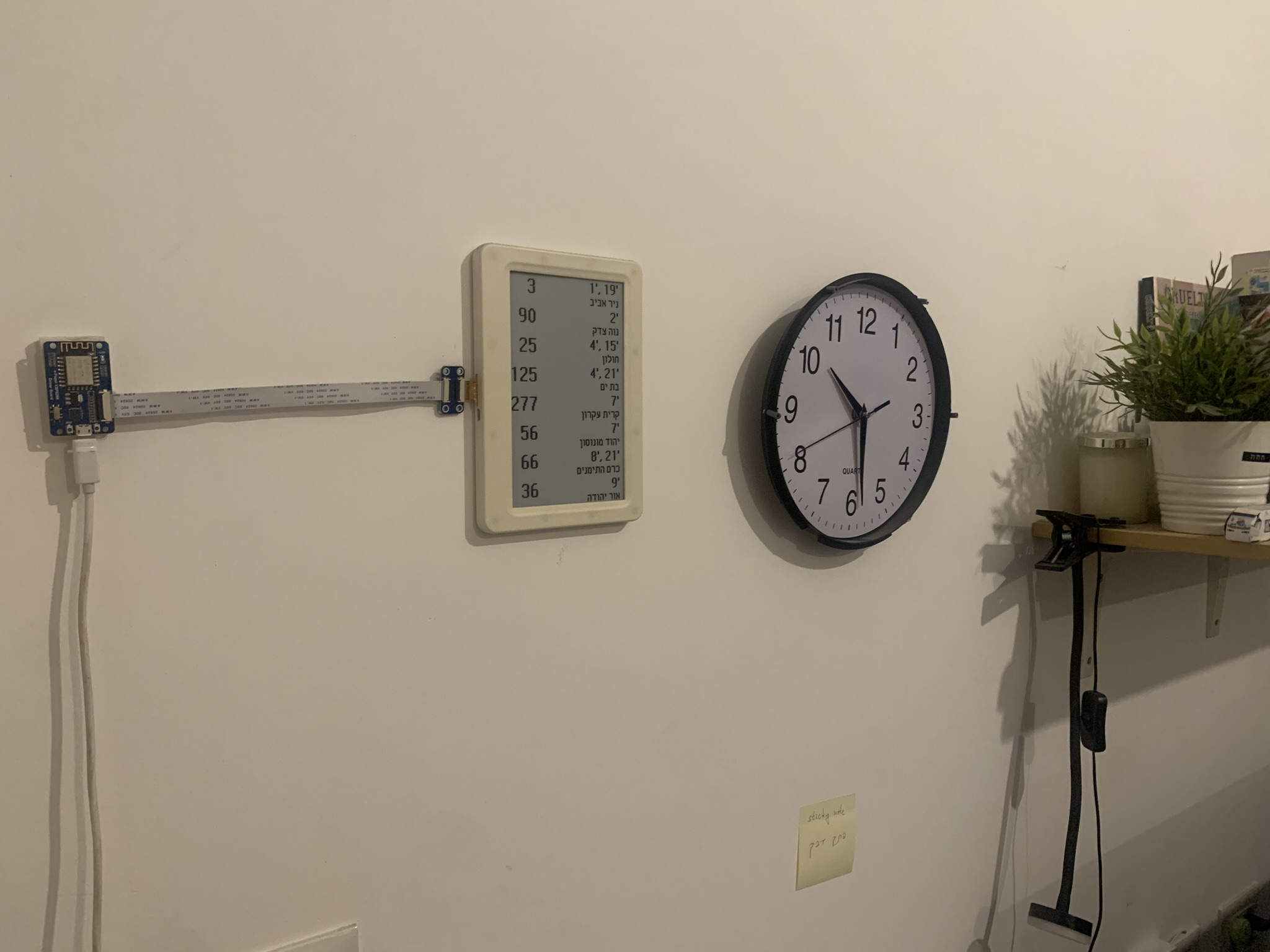 my living room wall. an e-paper display, framed in a white plastic and stuck to the wall, shows some bus departures in hebrew. it's connected by a ribbon cable to a bare circuitboard, also stuck to the wall. this setup is hung next to an analog clock and a shelf