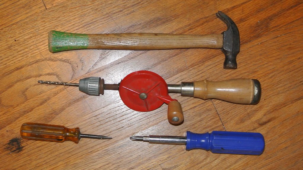 Claw hammer, hand drill and two screwdrivers laid out on a wooden floor