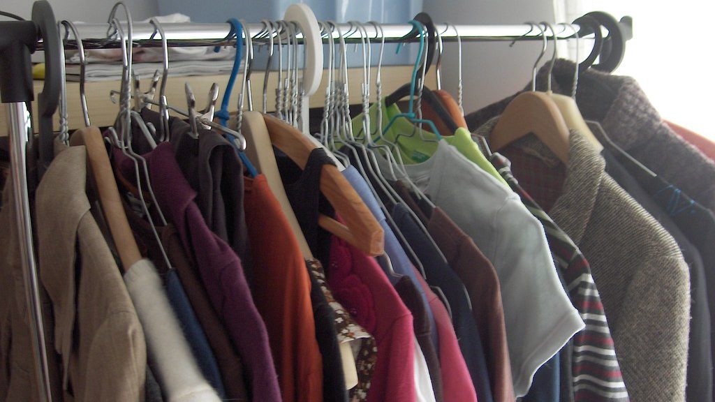 A rack of shirts, sweaters and jackets on wire and wooden hangers, with folded linens and a storage bin on the shelf behind