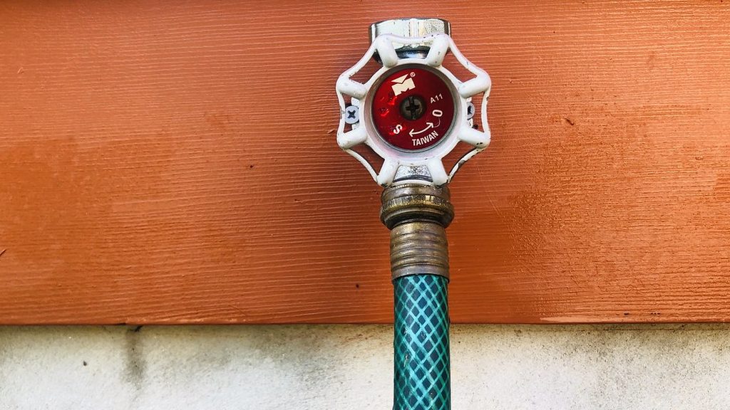 Outdoor hose faucet with a white handle on orange wood siding, a green garden hose still screwed onto it
