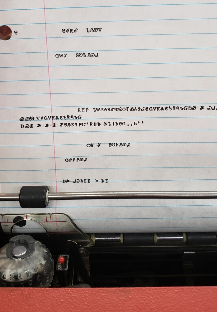 A sheet of lined paper in a typewriter with several lines of Cherokee text; the 3d printed typeball that made the text is visible in the lower left.