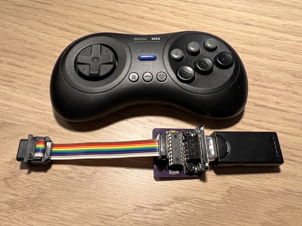 An 8BitDo M30 controller above an uncased circuit board populated with an 18-pin chip, a capacitor, a male DE-9 socket, and an IDC-connected ribbon cable terminated by a female DE-9 plug. An 8BitDo Retro receiver is plugged in to the adapter