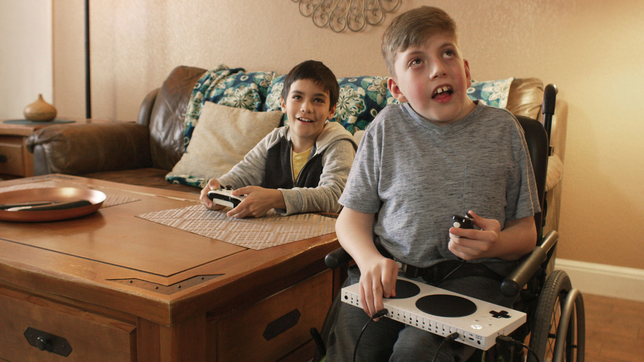 Duas crianças jogando videogame, uma delas está usando o controle padão do X-Box sentada no sofá e outra está usando o controle adaptado para pesssoa com deficiência e está sentada em uma cadeira de rodas