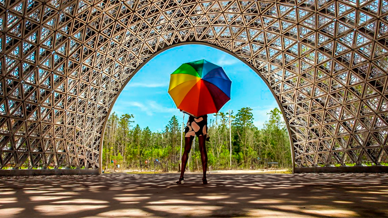 Pessoa com um guarda-chuva arco-íris está no meio de um túnel estruturado olhando para o céu azul e árvores