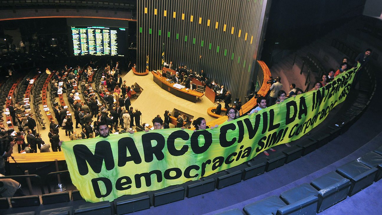 Câmara dos Deputados do Brasil. Em destaque, há um grupo de pessoas segurando uma grande faixa verde com letras pretas que diz 'MARCO CIVIL DA INTERNET - Democracia SIM! Corporações NÃO!'. Essa faixa está posicionada no andar superior do auditório, acima dos assentos dos legisladores. Ao fundo, vários parlamentares e funcionários ocupando as cadeiras e circulando pelo plenário. O ambiente é formal, com muitas cadeiras dispostas em fileiras semicirculares voltadas para uma mesa principal onde provavelmente estão posicionados os líderes ou o presidente da sessão.