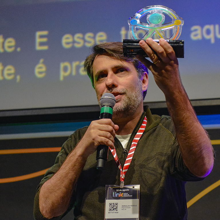 Reinaldo é um homem de pele branca, cabelos castanhos e barba grisalha e curta, vestido uma camisa com a cor escura e um crachá de identificação com um cordão listrado em vermelho e branco. Segura um troféu e fala ao microfone. O troféu que ele segura é translúcido e tem um círculo, dentro do círculo existe uma representação de uma pessoa os as pernas e os braços abertos.