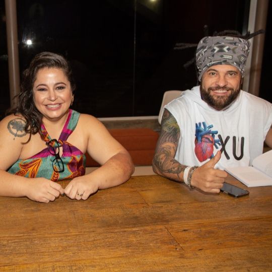 Duas pessoas cadeirantes, com os braços apoiados sobre a mesa. A Carolina é uma mulher branca, de cabelos longos e lisos, tem uma tatuagem no ombro direito, veste uma roupa estampada e as alças estão cruzadas no pescoço. O Billy é um homem pardo, usa dreadlocks e uma faixa grossa, cinza e estampada e tem os braços cobertos por tatuagens. Veste camisa branca sem mangas.