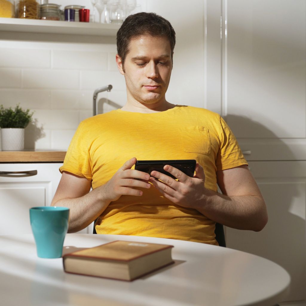 Homem de camisa amarela, sentado, segurando um celular com as duas mãos. No ambiente: na sua frente uma mesa, acima dela uma caneca esverdeada e um livro fechado, ao seu lado uma cadeira vazia. Ao fundo uma bancada com uma pia e fogão, alguns vasos de plantas e potes com temperos em cima de uma prateleira. 