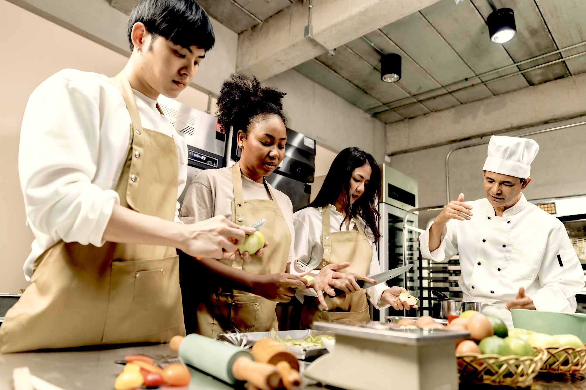 Four people in a cooking class, each peeling fruit with a different implement while an instructor guides them