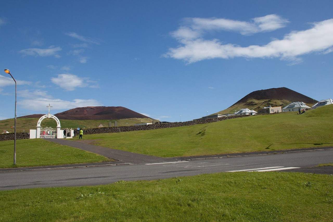 Heimaey / Vestmannaeyjar: Eldfell and Helgafell volcanic cones above cemetery gate, day, blue sky