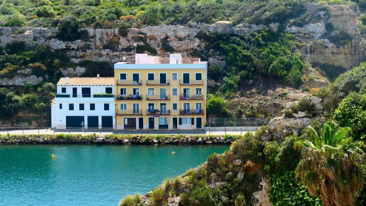 Mahón harbour cliff houses under limestone cliff, Menorca, day, turquoise water, blue sky