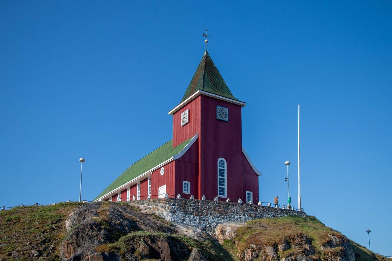 Sisimiut Bethel Church (red wooden church with green roof and clock tower) on rocky hill, day, solid blue sky