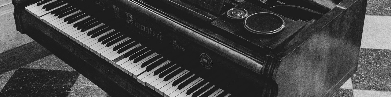 A black and white image of an old piano. The piano is an upright model, with the keys on the right side of the image. The piano is sitting on a tiled floor. There is a small round object on the top of the piano.