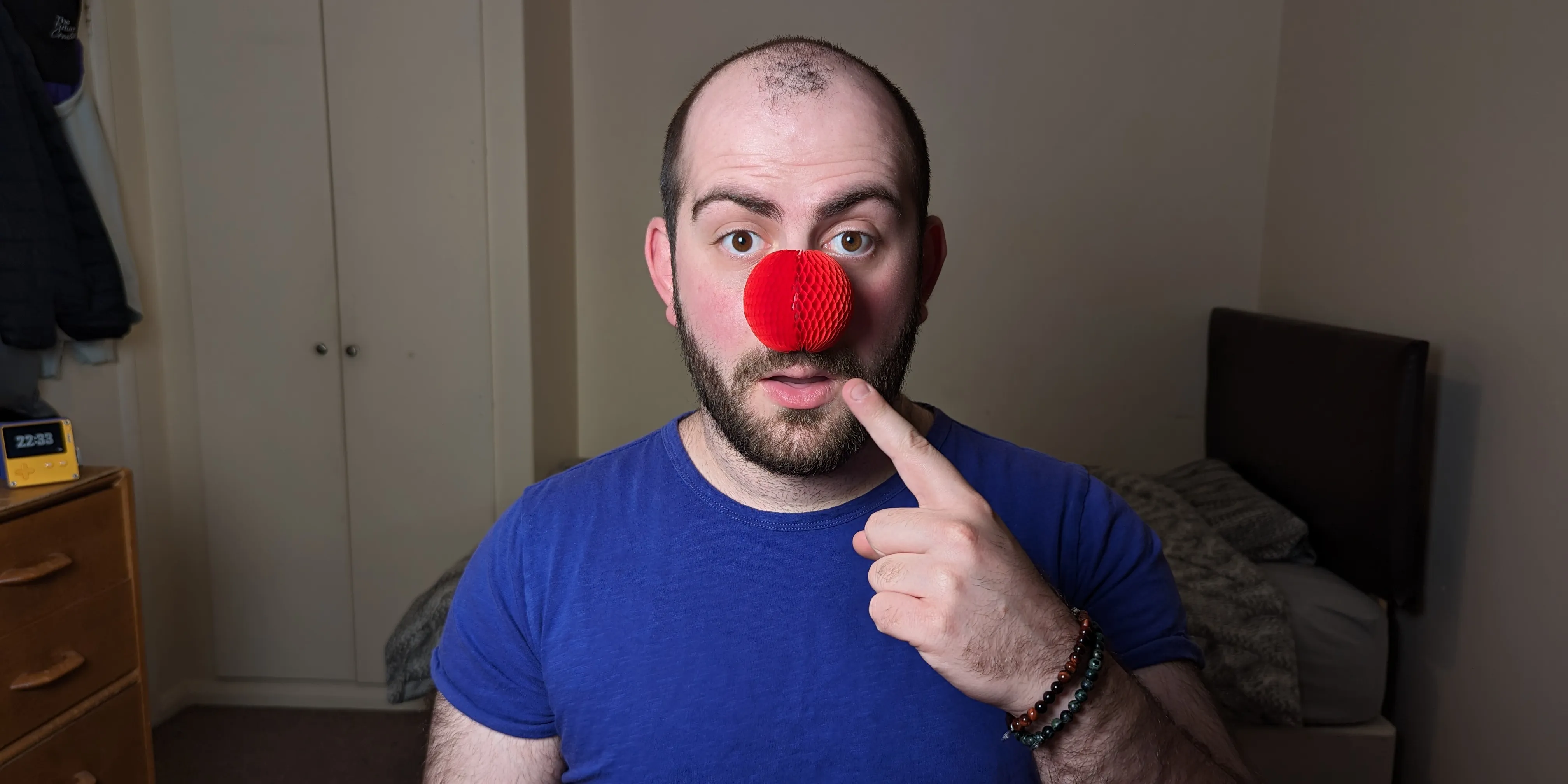 A picture of George wearing a red nose whilst pointing to it with a surprised expression on his face.