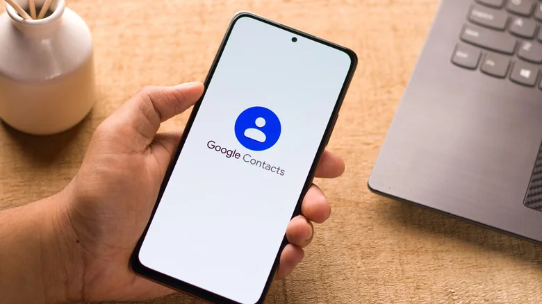 Google Contacts text and logo on a smartphone, held near a laptop.