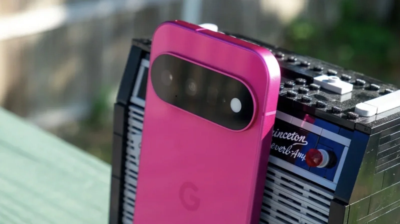 Photo of a hot pink Google Pixel 11 smartphone leaning against an Lego build of an audio amp in an outdoor setting.