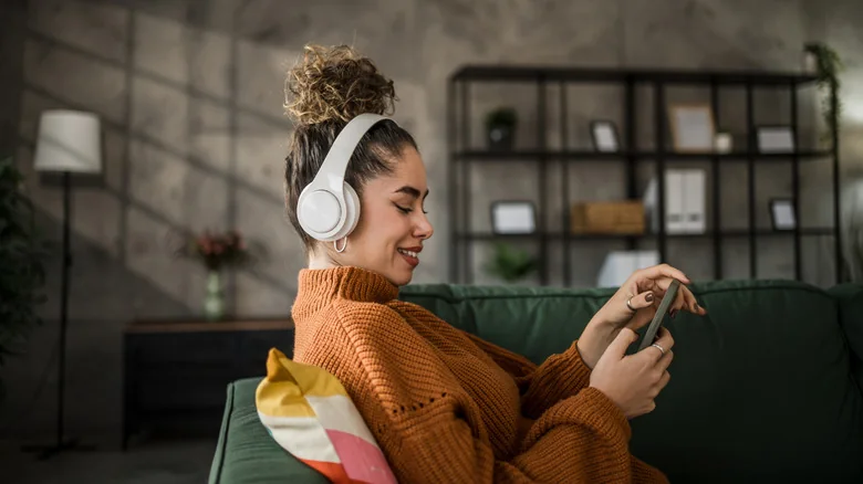 A woman wearing white headphones and an orange sweater smiles as she chooses music on her phone.