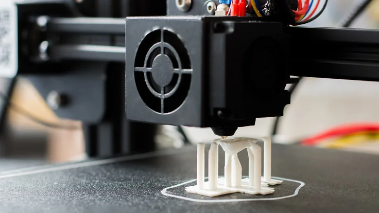 Close-up of a 3D printer nozzle printing a small white object on a black build plate.