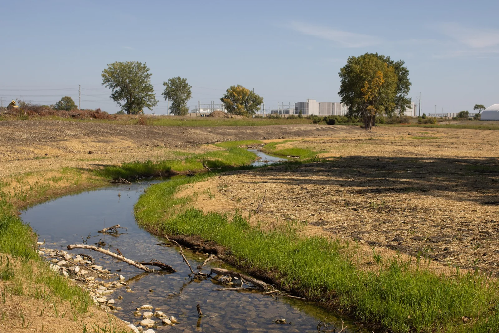 A creek seen flowing through a field with buildings in the background.