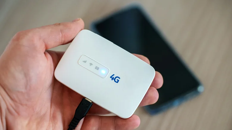 Hand holding a wireless 4G hotspot device above a blurred smartphone in the background.