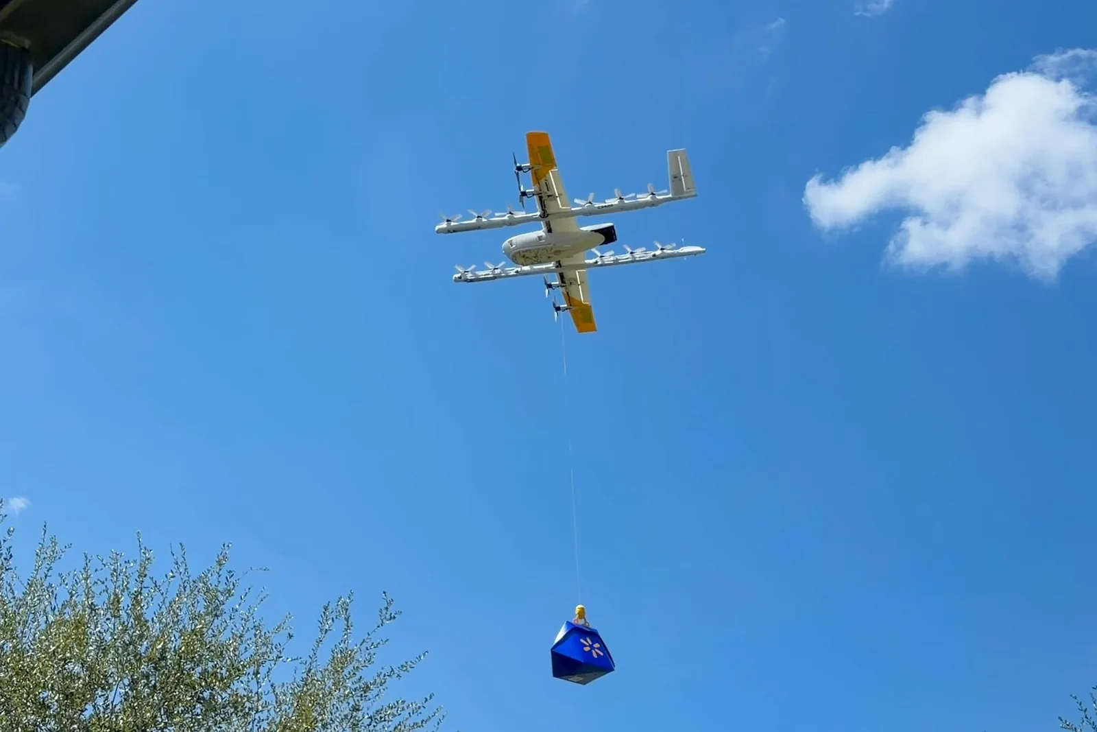 Fixed-wing drone with a bunch of propellers lowering a blue Walmart doggy bag.