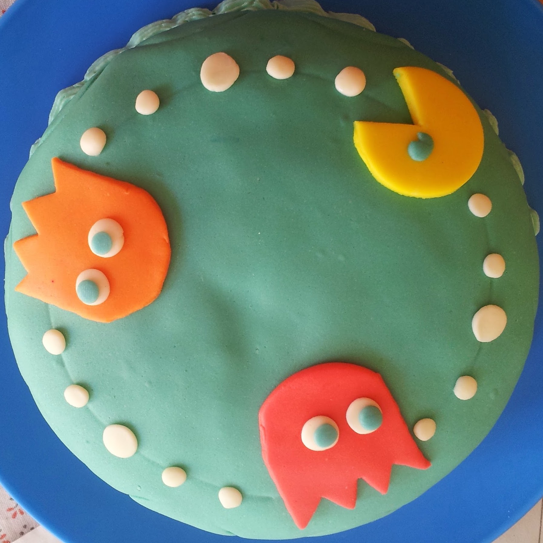The typical photo JorgeArtware uses everywhere is a jungle-green cake with pacman thematic for the edible decorations arranged in a circle: there's the famous yellow character with jungle-green eyes, fliped upside-down at the top right which makes it seem as if it was defying gravity, because the center of the circle is meant to be the sky, and there's two ghosts: Blinky(red) & Clyde(orange), the red one at the bottom facing to the right and the orange at the left facing down, every character is separated by 5 white dots, the one in the middle is a big yummy one every time. 