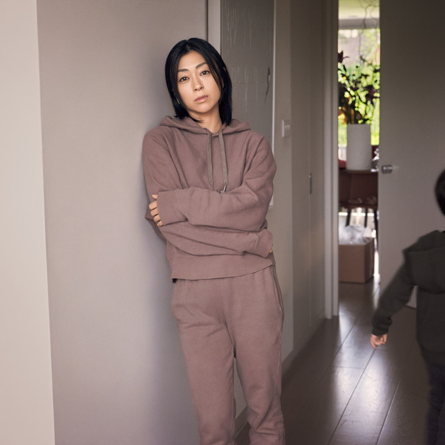 Album art for BADモード album with Utada Hikaru in her brown sweats, standing in a hallway in what seems to be her home, against a while wall, with a child at the edge of the frame running away from her