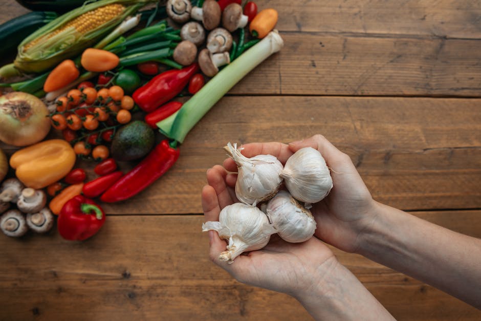 Hands holding garlic with a variety of fresh vegetables on a wooden table. Overhead shot.