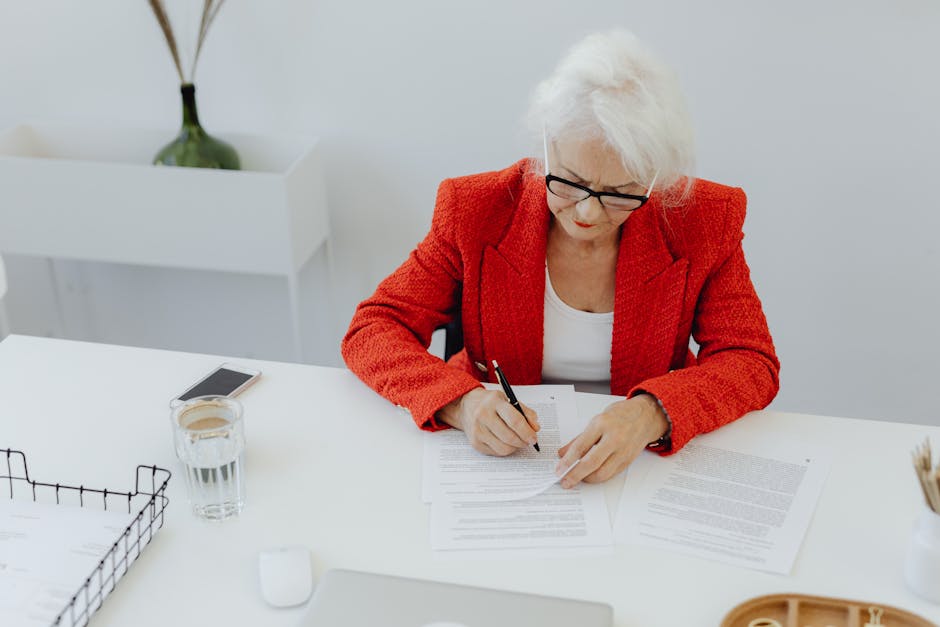 Elderly woman in red jacket writing at a tidy desk in a modern office setting.