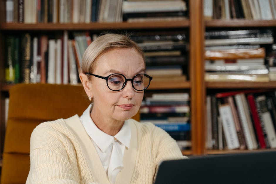 A senior woman wearing glasses focusing on her laptop in a cozy library environment.