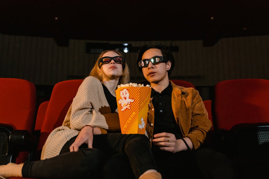 Two adults wearing 3D glasses watching a movie with popcorn in a cinema setting.