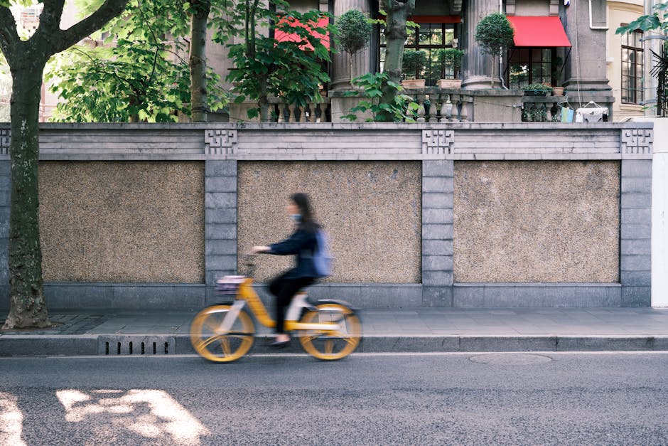 Blurred woman cycling past urban sidewalk and stone wall with greenery.