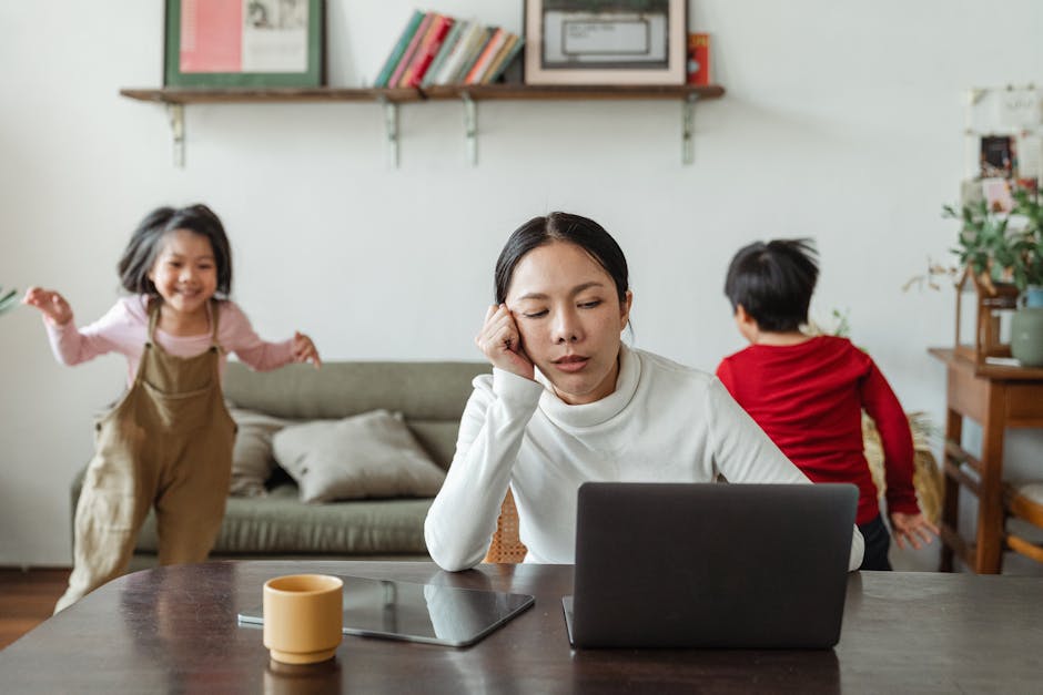 Exhausted young Asian female working distantly from home with happy noisy children playing around