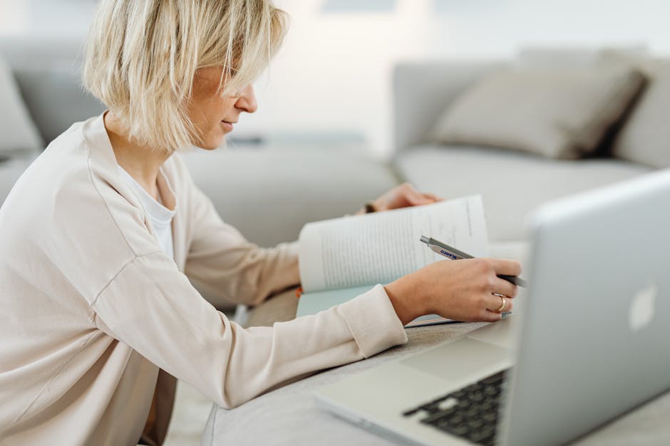 Woman with blonde hair intently studying with a book and laptop in a cozy indoor setting.