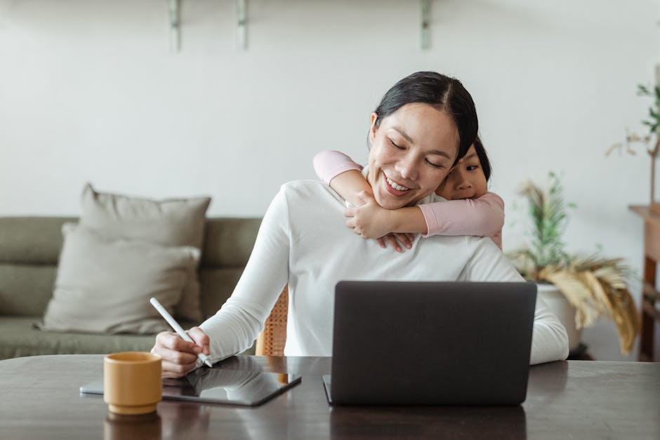 Positive Asian mother working online using laptop and tablet and cute small daughter seeking attention and tenderly embracing mom from behind