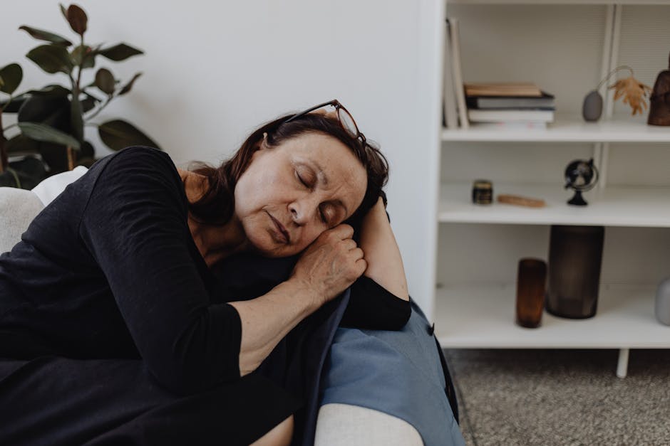 Senior woman in casual attire sleeping on a sofa with a serene expression indoors.