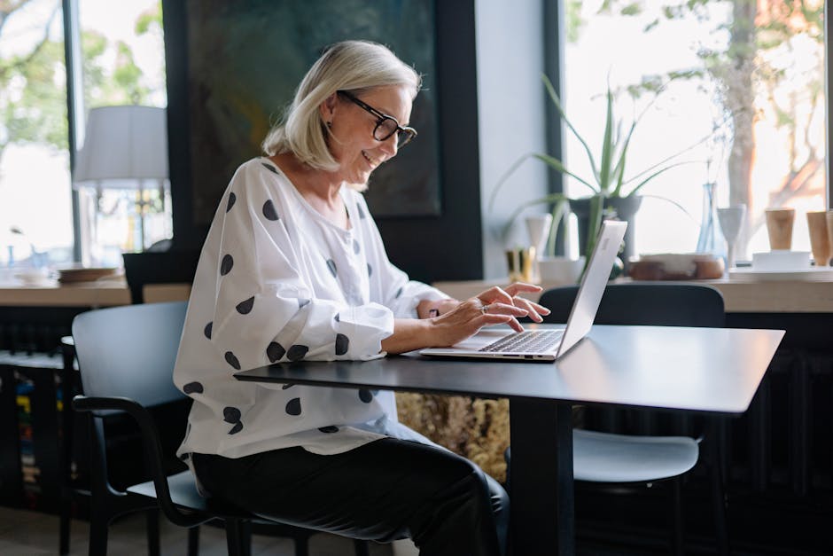 Middle-aged woman in polka dot blouse working on a laptop by a window.