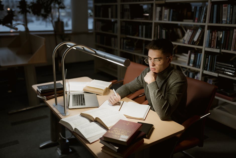 A young adult studying at a library desk with books and laptop under warm lighting.