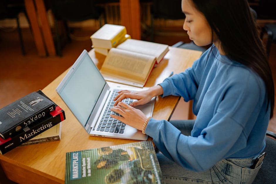 Young woman typing on a laptop surrounded by textbooks in a library setting.