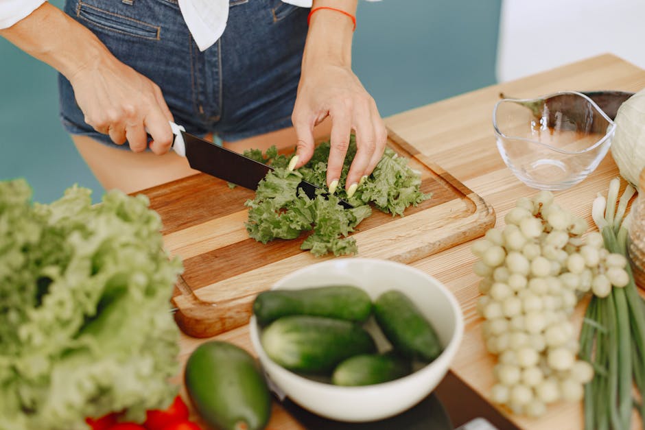Hands chopping leafy greens on a wooden board with fresh vegetables for a healthy meal preparation.