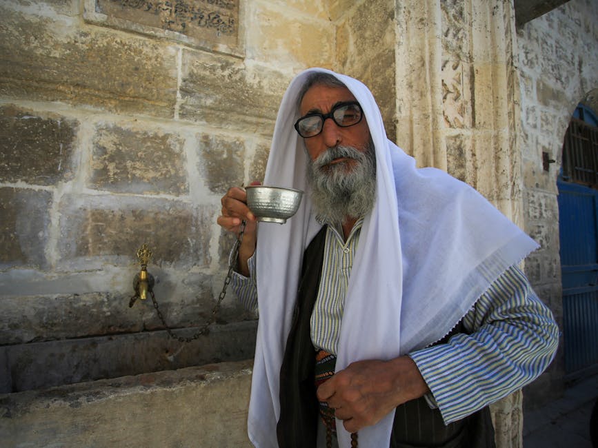 Elderly Middle Eastern man with glasses holding a metal cup near traditional stone wall.