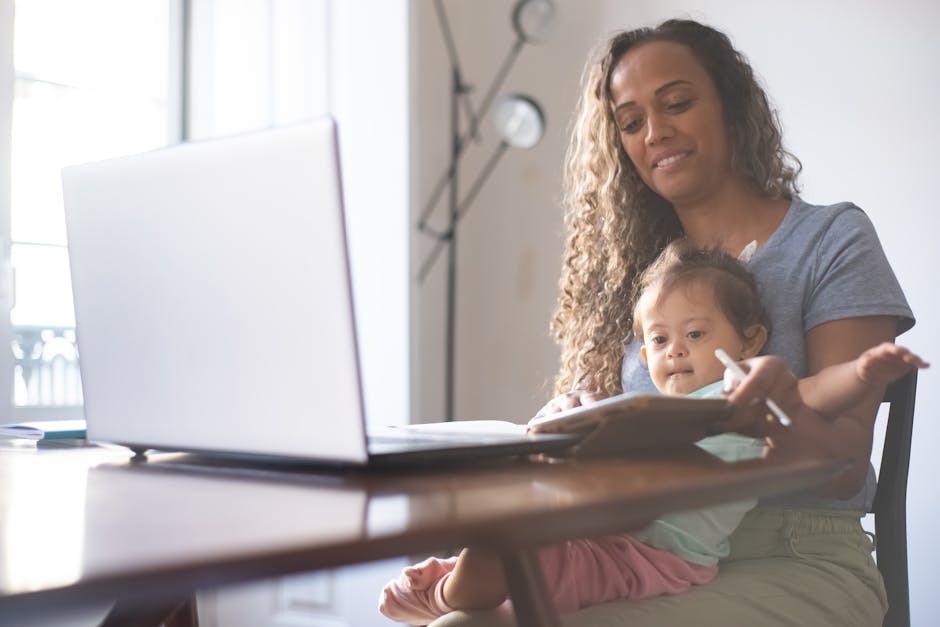 A mother multitasks at a desk with a laptop and her child in a bright home setting.