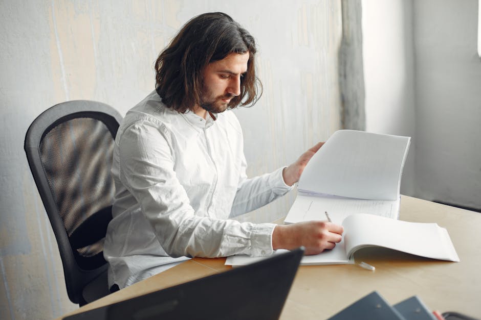 A young man working remotely, reviewing paperwork at a desk indoors.